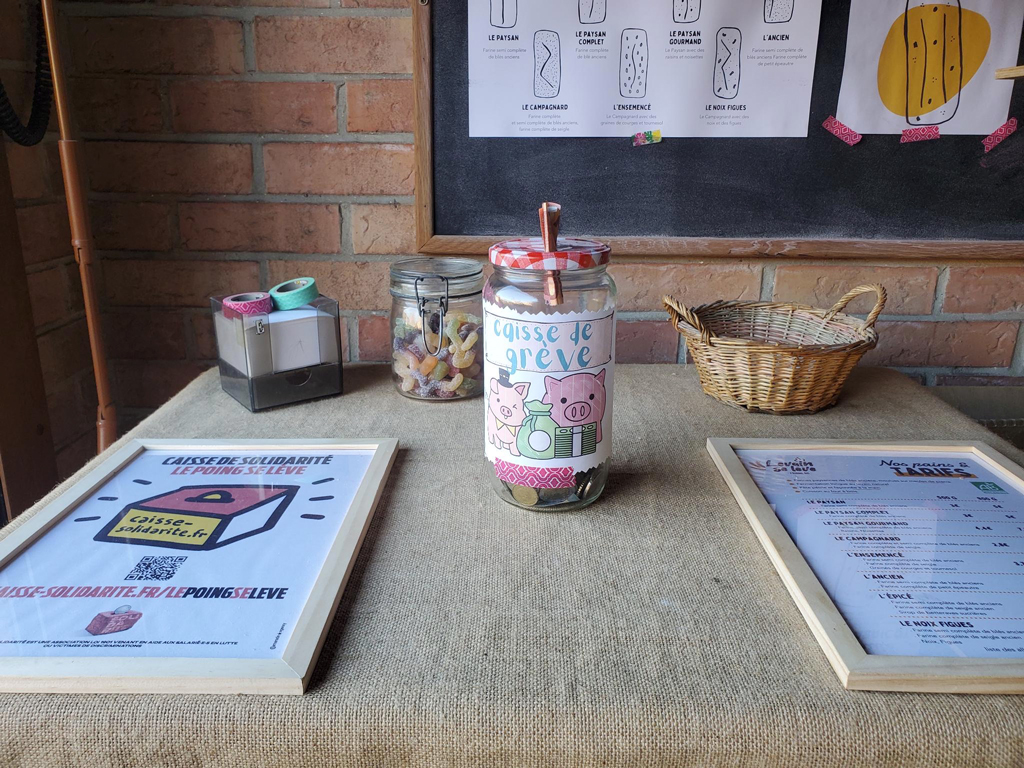 Bocal en verre étiqueté « caisse de grève » servant d’urne à dons sur le comptoir d’une boulangerie, à côté d’une affiche « Caisse de solidarité — le poing se lève ».
